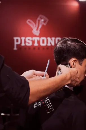 Barbeiro fazendo o acabamento com navalha em frente à parede vermelha com o logo Pistons